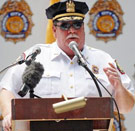 It's bright in here, okay? Now sit down, shut up, and next question. Police Chief Commissioner standing at podium giving a speech