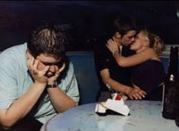 Guy sitting lonely at a table while his friend makes out with a girl.