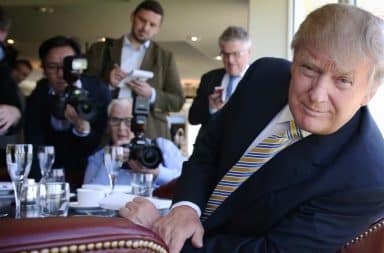 Donald Trump smirks at table while being photographed by media