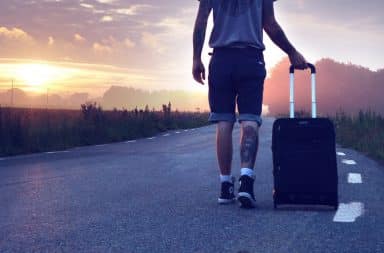 Man wearing jean shorts walking his suitcase down the road into the sunset