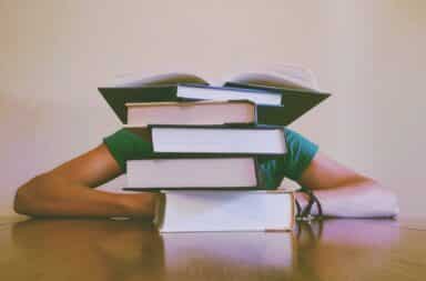 Person with a pile of books in front of face at a desk